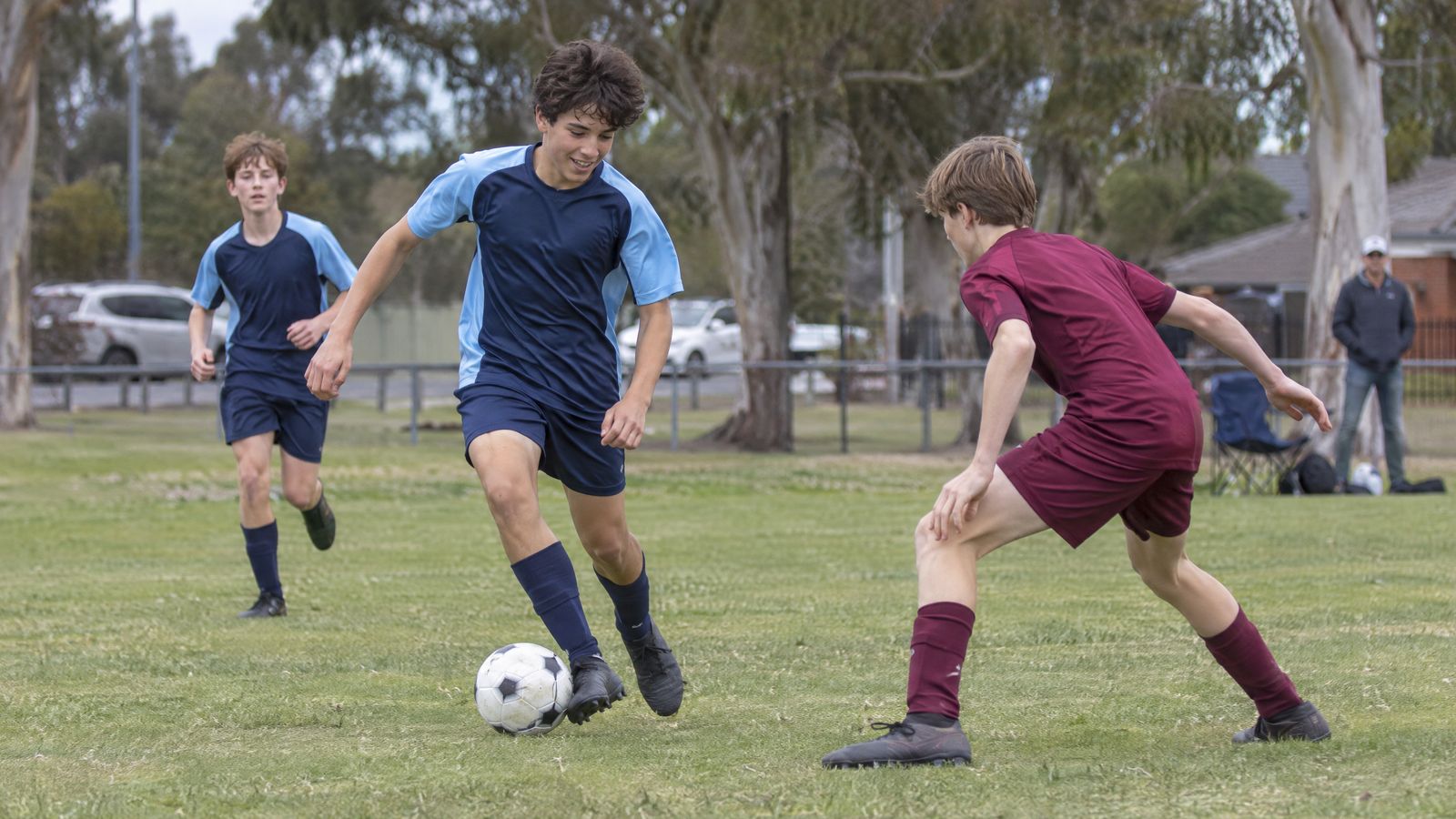 A teenage footballer in a blue kit dribbling past a defender in maroon on a suburban oval