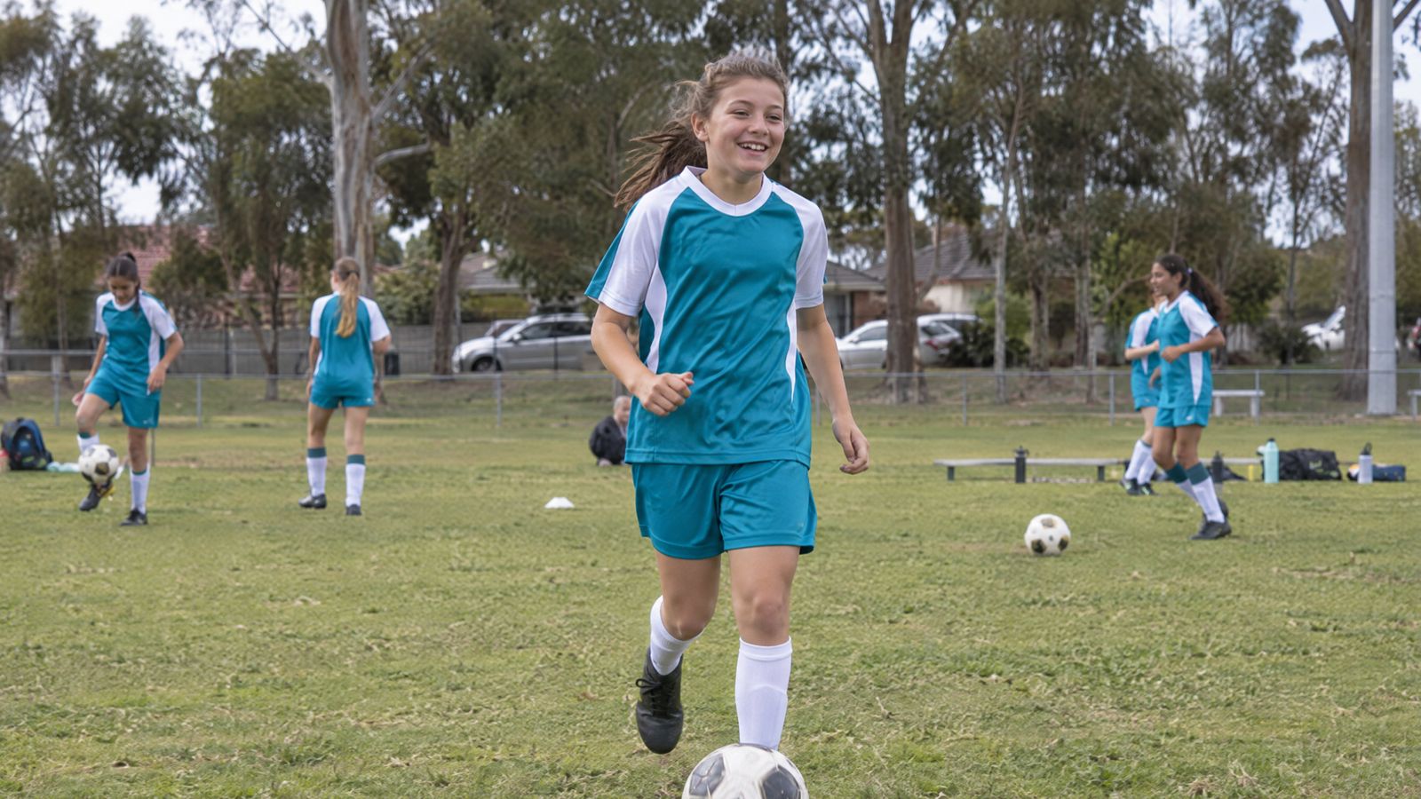 A smiling girl in a teal soccer kit jogging onto a suburban oval with a ball