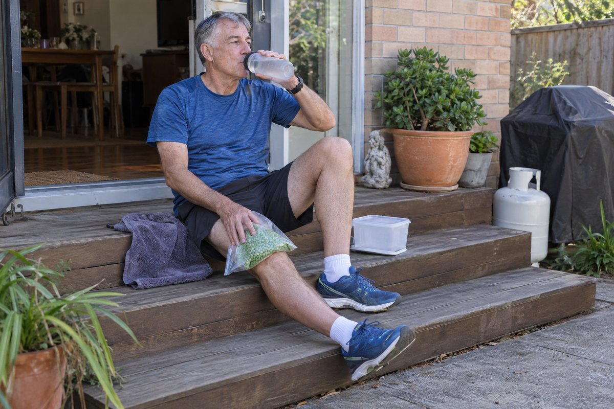 A grey-haired runner sitting on his back steps after a run, a bag of frozen peas on his knee, drinking water