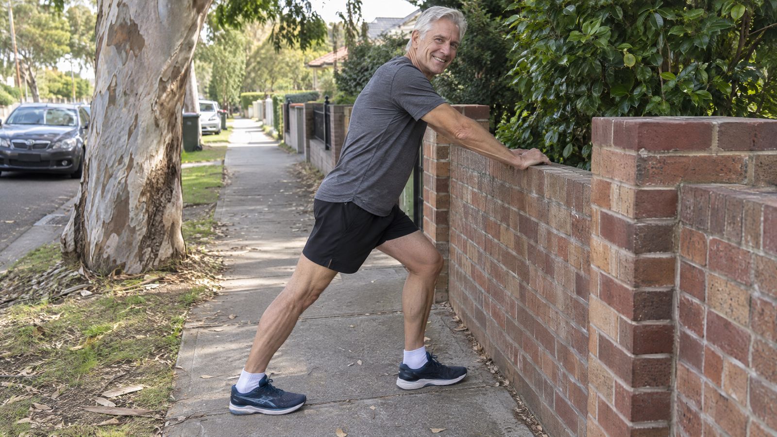 A grey-haired runner stretching his calf against a brick fence on a suburban footpath, smiling