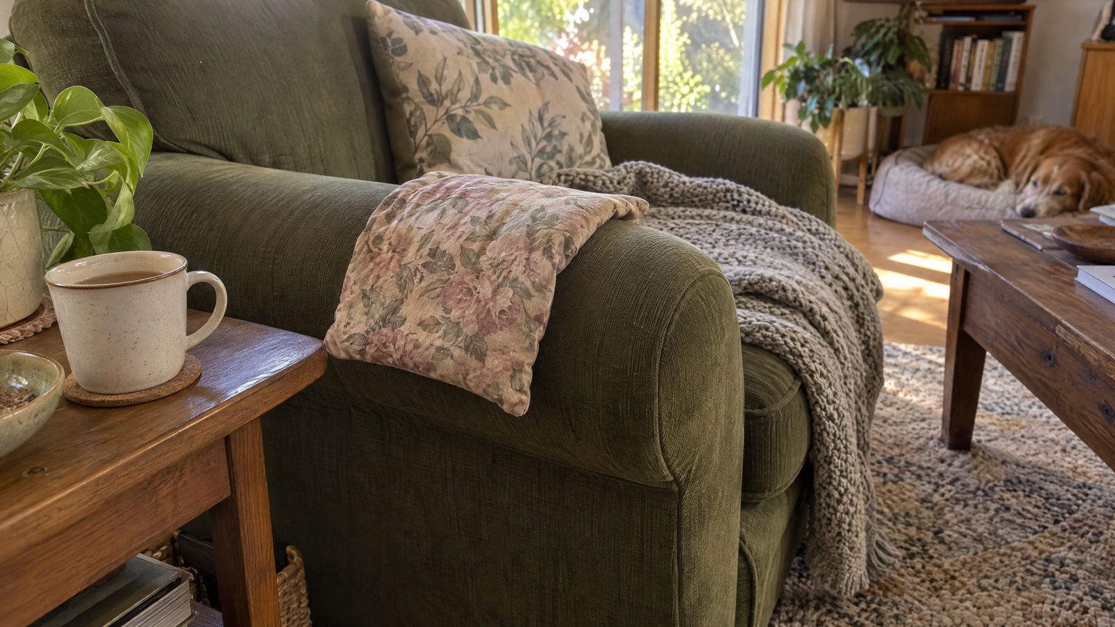 A floral wheat bag draped over the arm of an olive armchair, a cup of tea on the side table